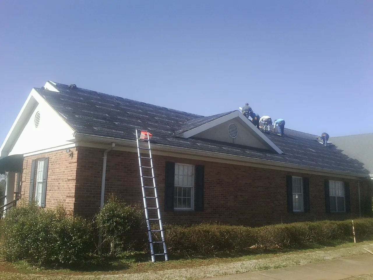 Crew tearing shingles off a brick ranch roof with a ladder set — Metal Roof Repair work by our crew in Waller