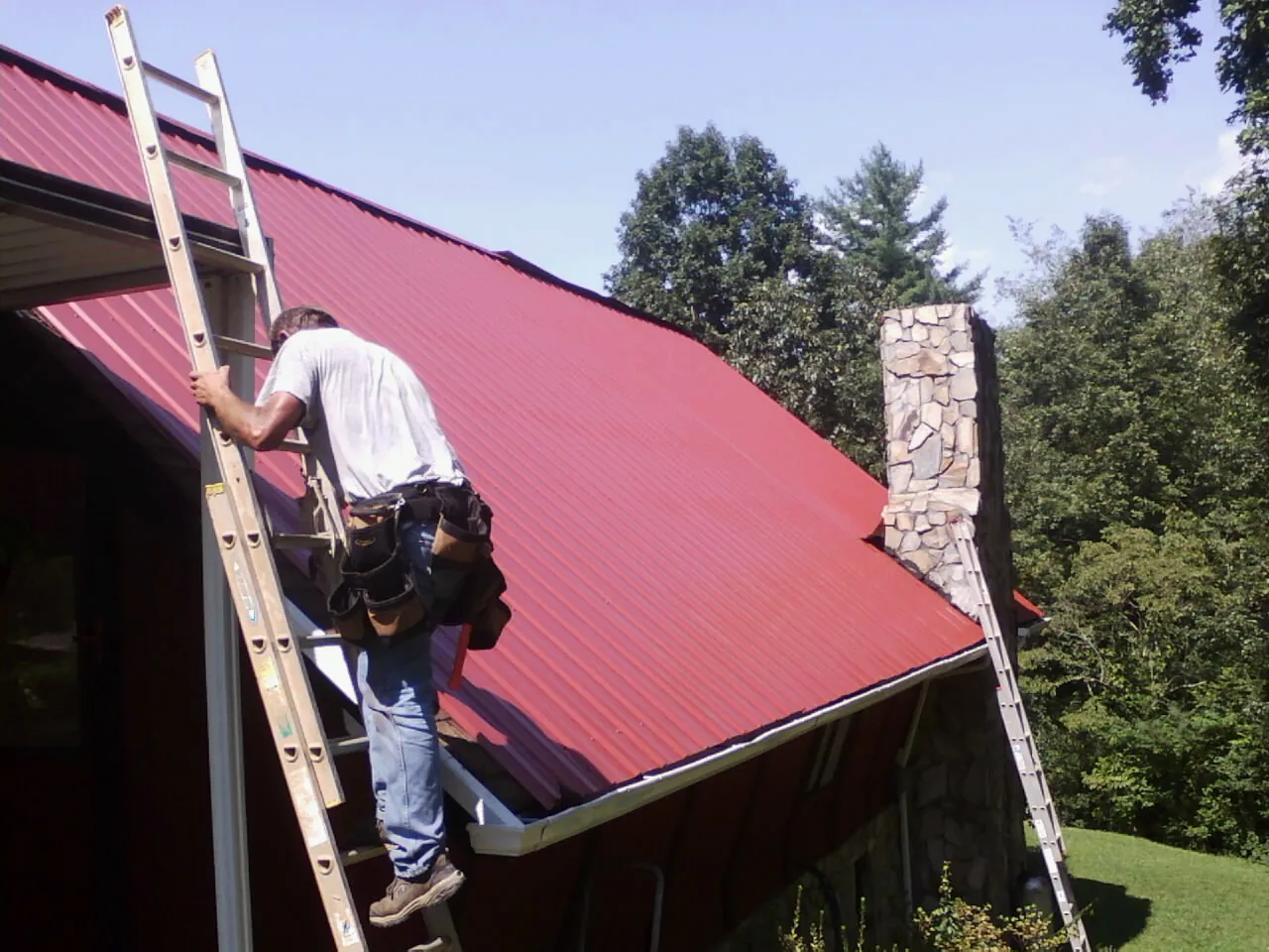 Roofer on a ladder beside a red metal roof and stone chimney — Soffit & Fascia Repair by Roofing Brothers in Waller